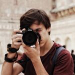 man in red and black crew neck t-shirt holding black nikon dslr camera