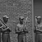 grayscale photo of 2 men standing near brick wall