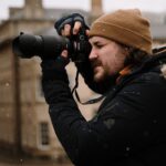 man in black jacket and brown knit cap holding black dslr camera