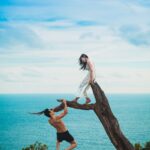 woman sitting on tree trunk with man holding on branch near sea under white clouds during daytime
