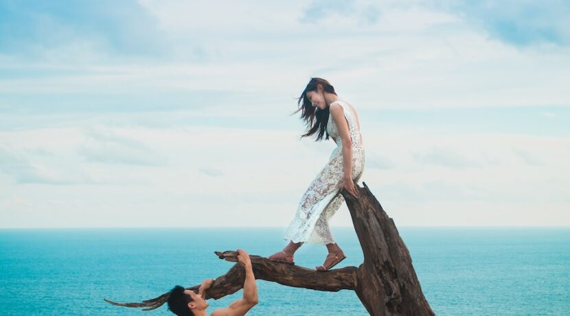 woman sitting on tree trunk with man holding on branch near sea under white clouds during daytime