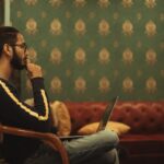 man sitting on chair while using laptop on his lap near sofa inside room