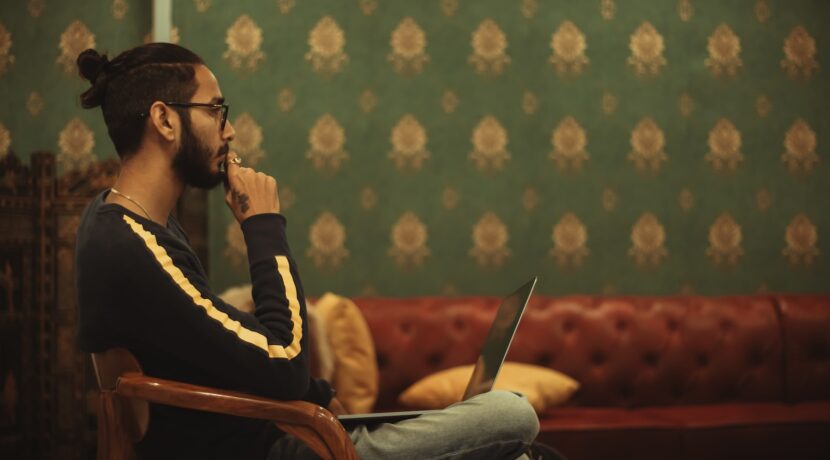 man sitting on chair while using laptop on his lap near sofa inside room