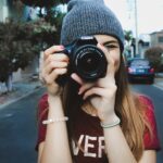 woman wearing red t-shirt and gray knitted cap standing on concrete road using Canon bridge camera during daytime