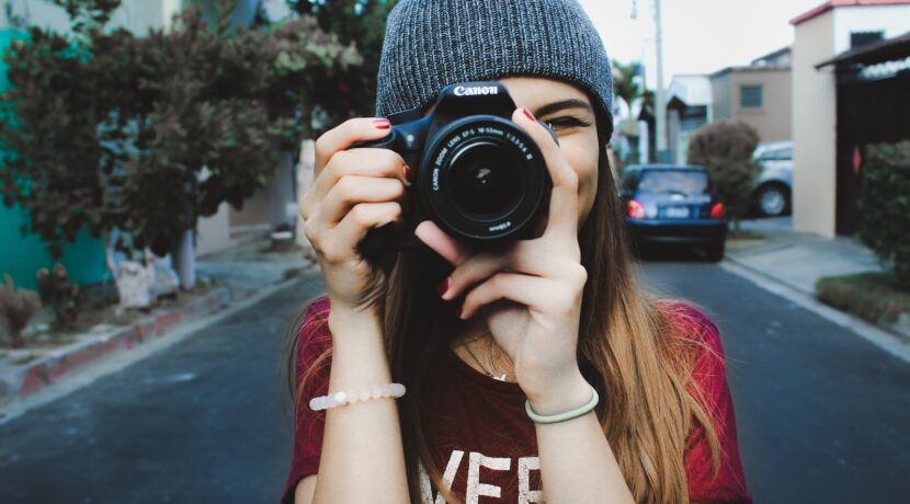 woman wearing red t-shirt and gray knitted cap standing on concrete road using Canon bridge camera during daytime