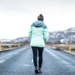 woman wearing teal jacket and black pants walking on concrete road