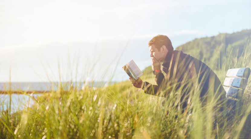 man reading book on beach near lake during daytime