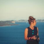 woman wearing black V-neck sleeveless dress holding DSLR camera in front of sea