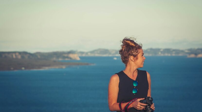woman wearing black V-neck sleeveless dress holding DSLR camera in front of sea